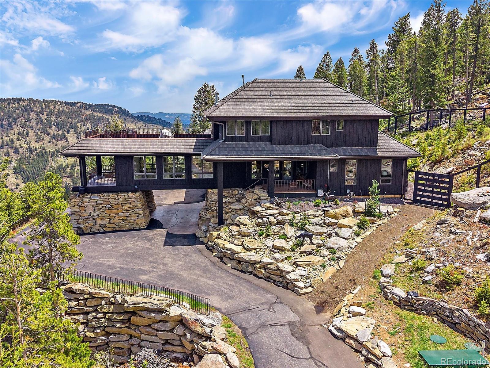 Dark-sided mountain home with stone retaining walls and a paved driveway, set among pines with mountain views.