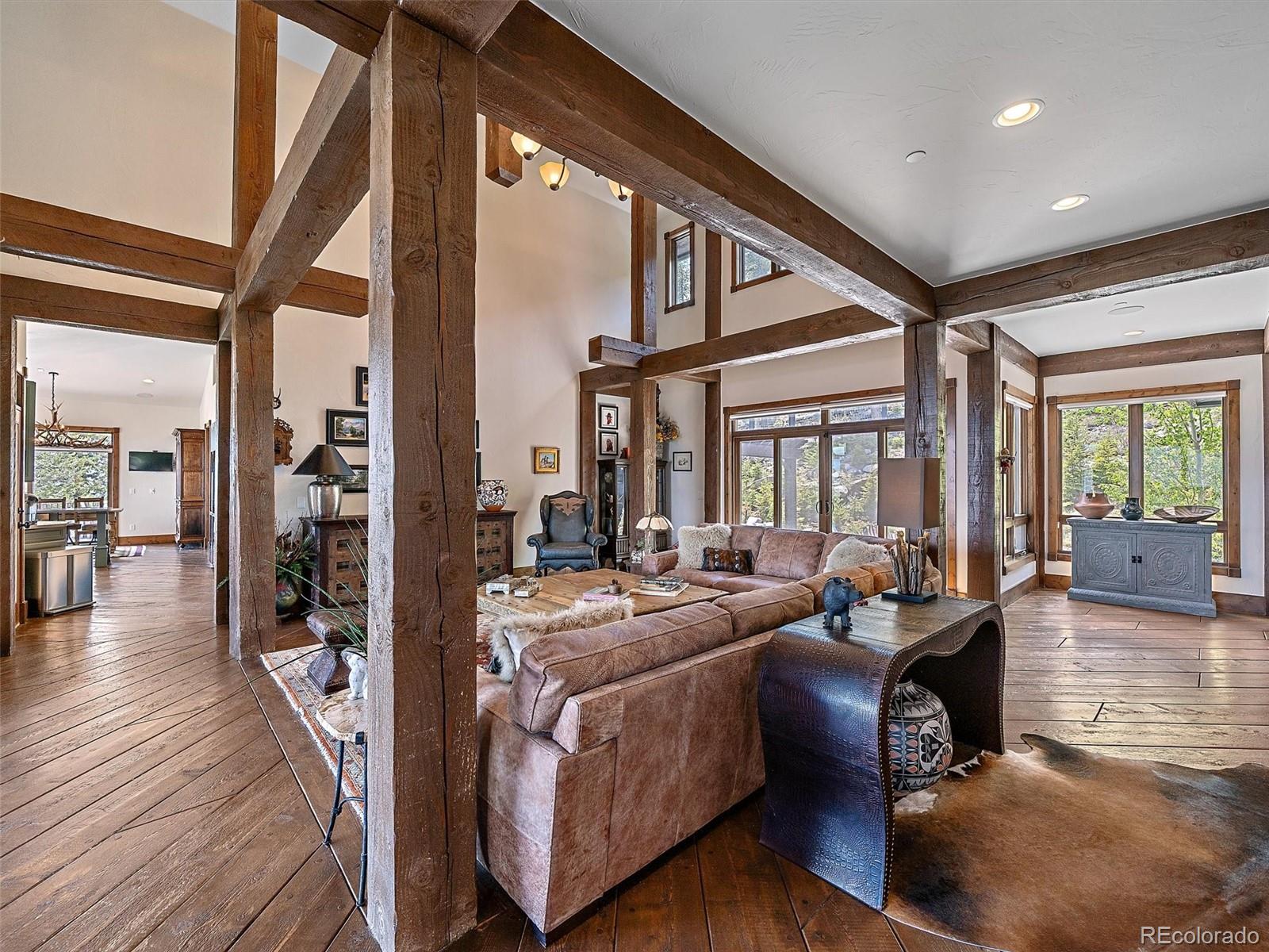 Living room with exposed timber beams, vaulted ceiling, wood floors, and large windows overlooking trees.