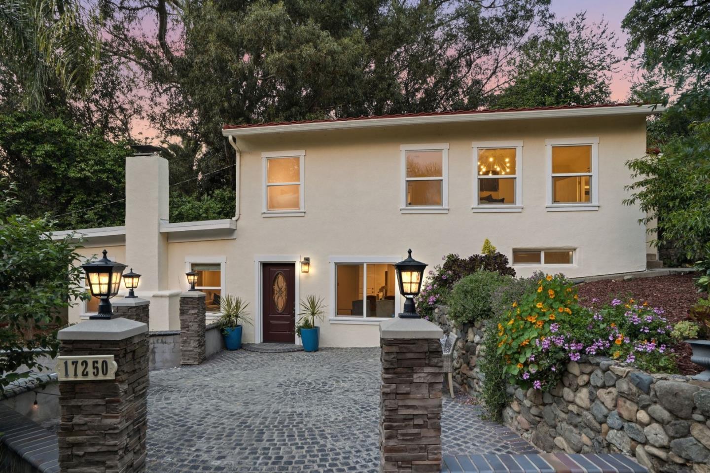 Two-story cream stucco home with a cobblestone driveway, stone pillars, and flowering landscaping at dusk.