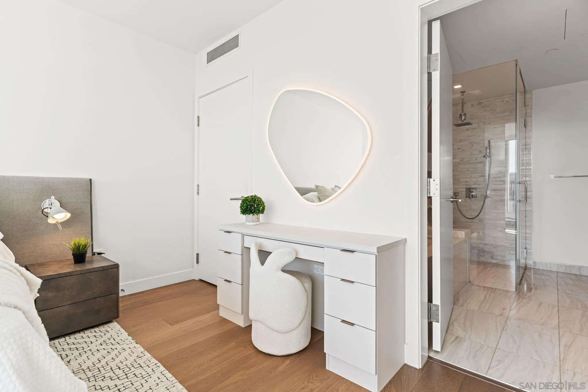 Bedroom with a white desk and illuminated mirror beside an open doorway to a glass shower.