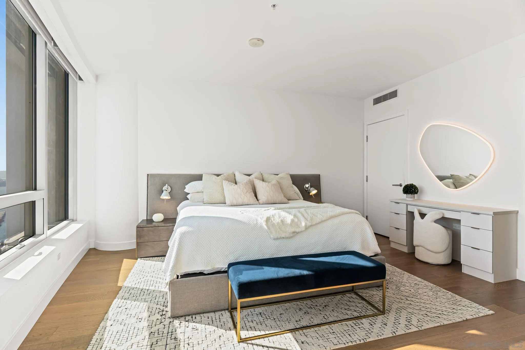 Bright bedroom with a white-dressed bed, navy bench, and illuminated mirror beside large windows.