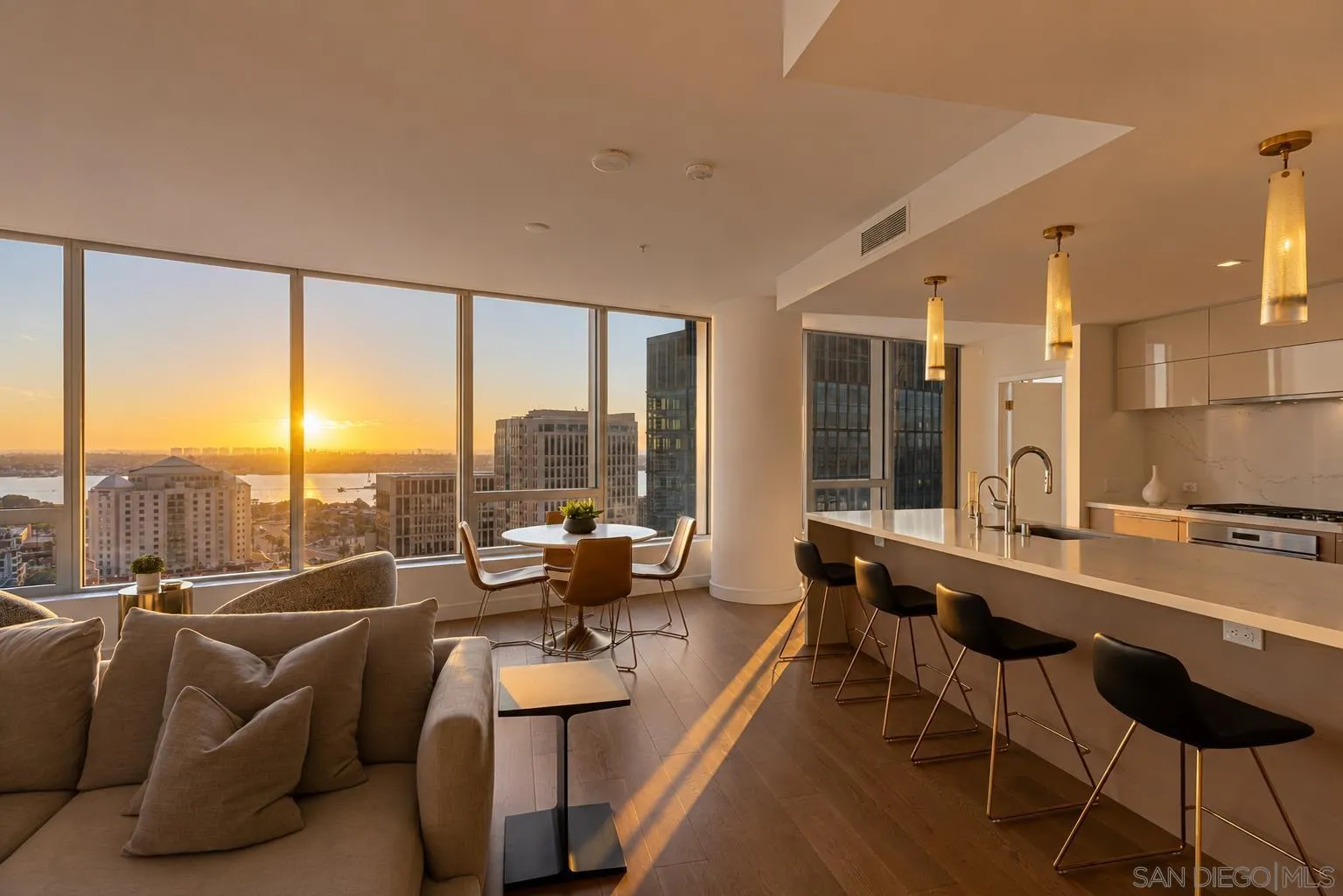 Furnished high-rise living room and kitchen with floor-to-ceiling windows overlooking downtown San Diego at sunset.
