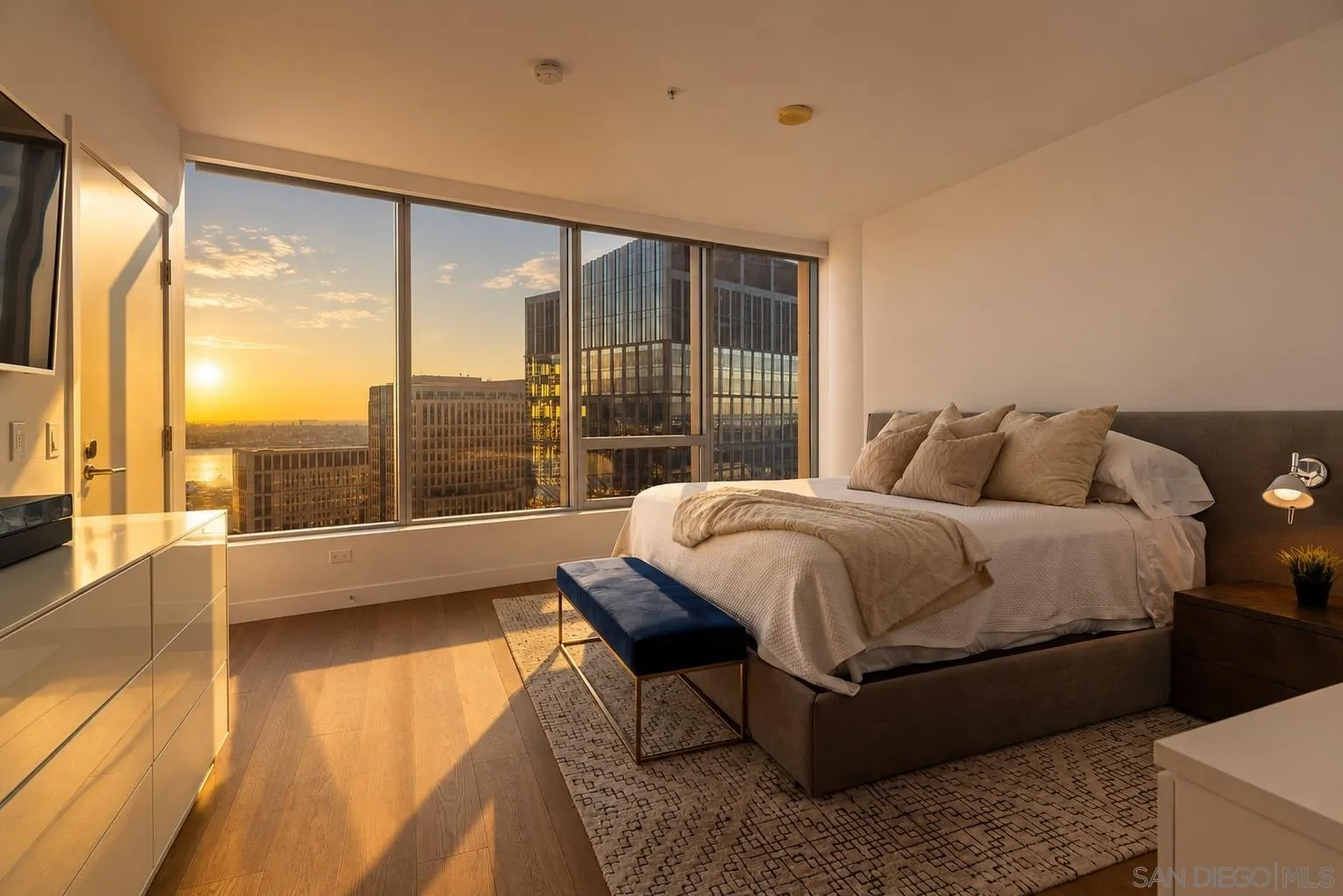 Furnished high-rise bedroom with floor-to-ceiling windows overlooking downtown and the water at sunset.