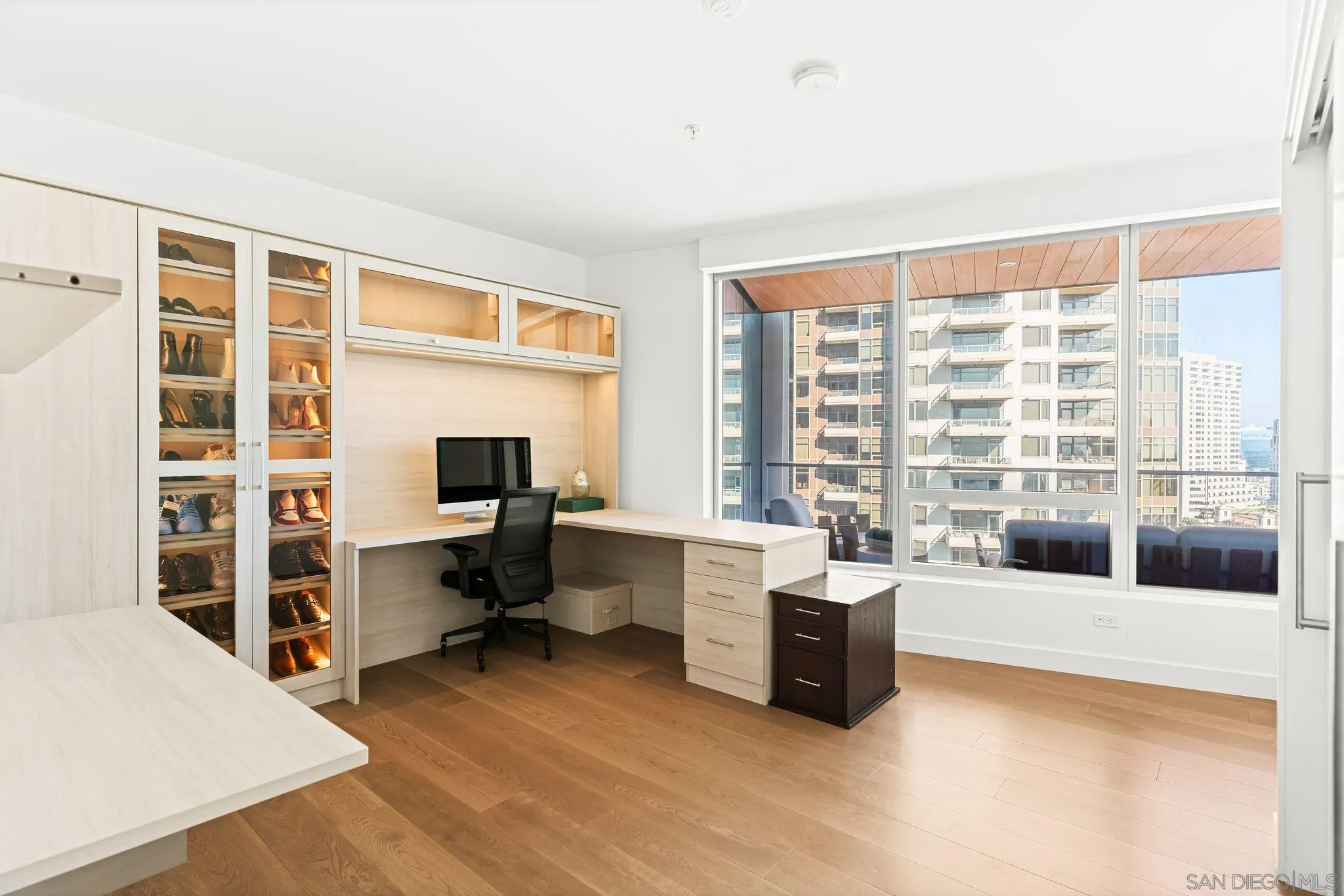 Home office with built-in desk and shoe storage beside large windows overlooking downtown buildings.