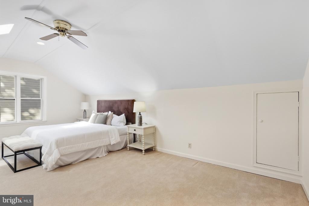 Staged bedroom with a white bed beneath a sloped ceiling and a ceiling fan.