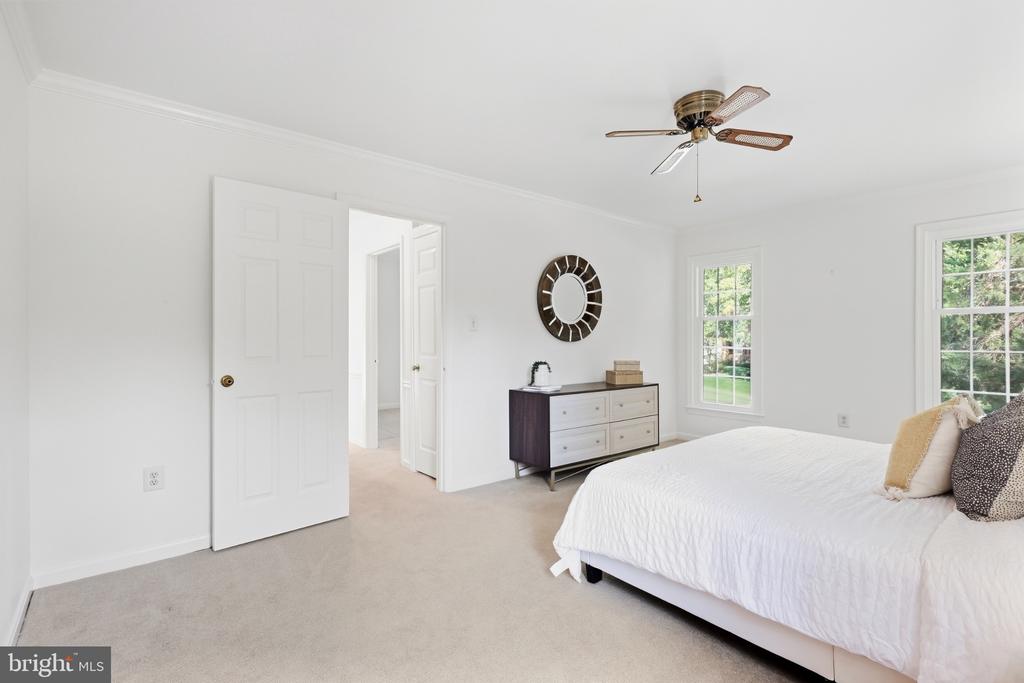Furnished bedroom with a white bed, beige carpet, ceiling fan, and windows overlooking greenery.