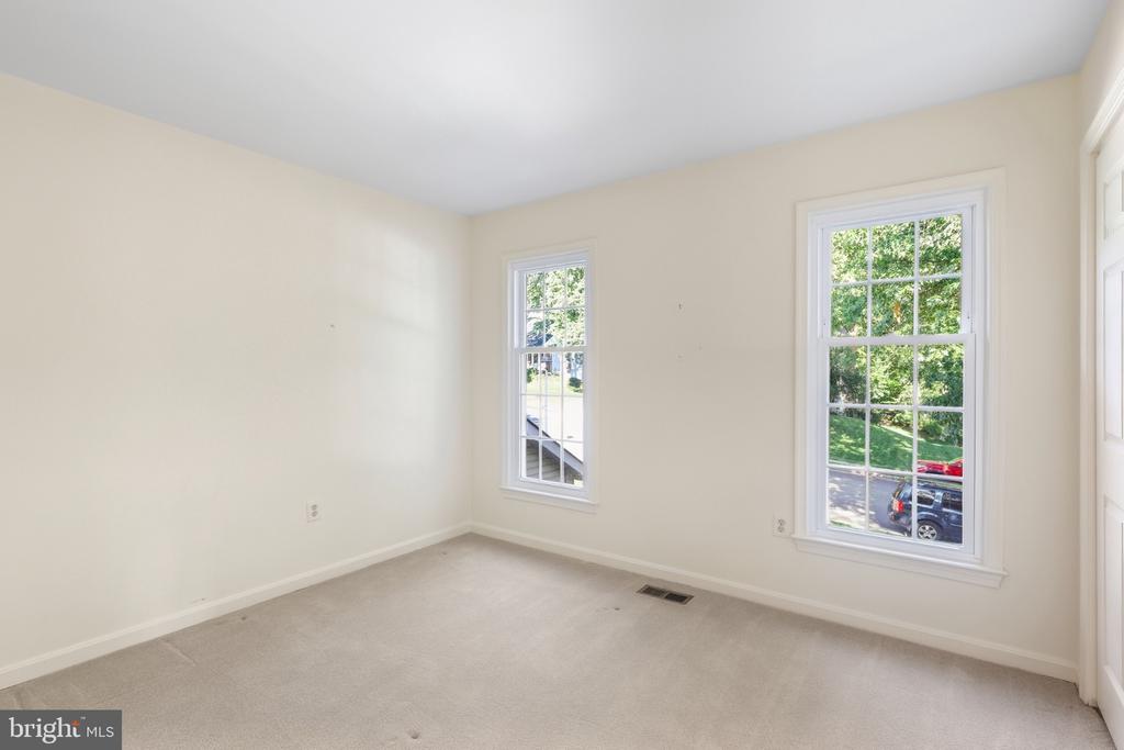 Empty carpeted room with cream walls and two tall windows overlooking a leafy street.