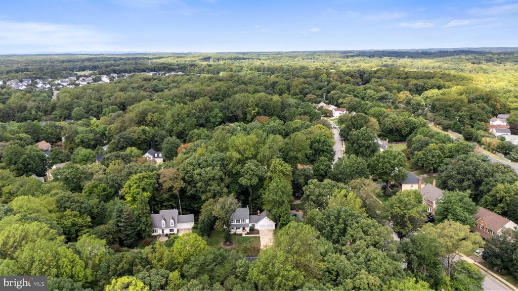 Aerial view of a tree-covered neighborhood, with a white home near the foreground and wooded hills beyond.