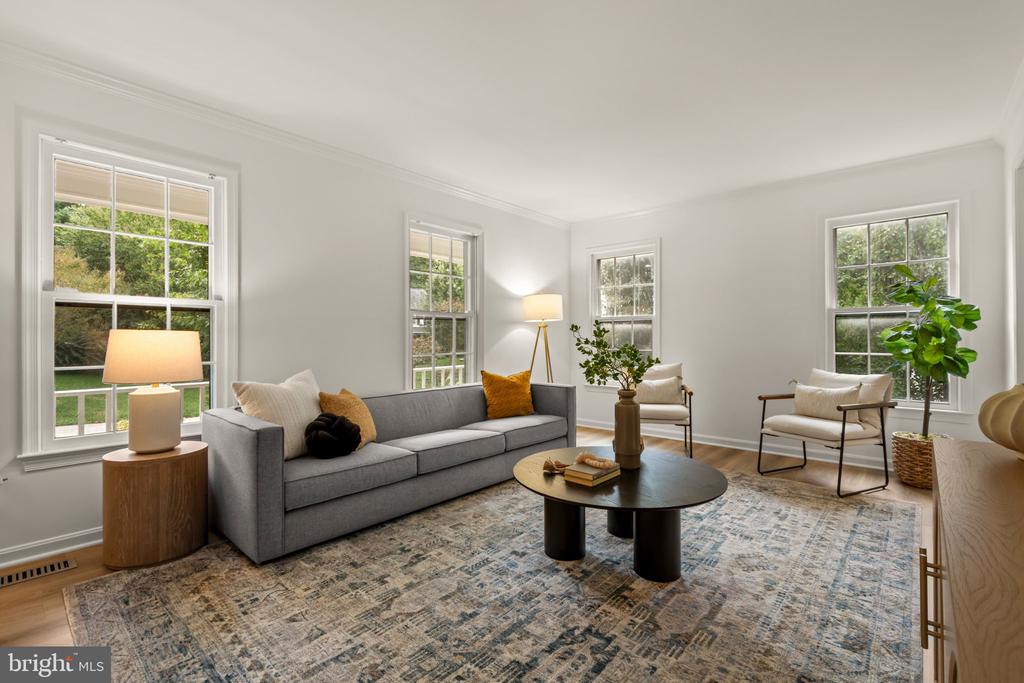 Staged living room with a gray sofa, round coffee table, patterned rug, and windows overlooking greenery.