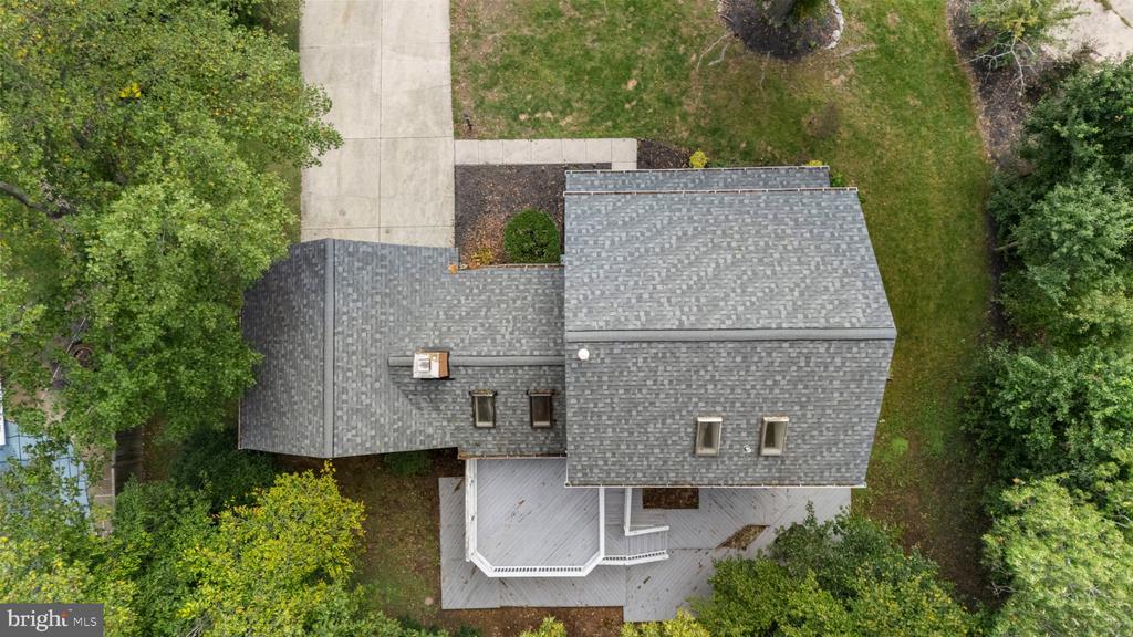 Aerial view of a gray-roofed house with a rear deck, concrete driveway, and leafy yard.