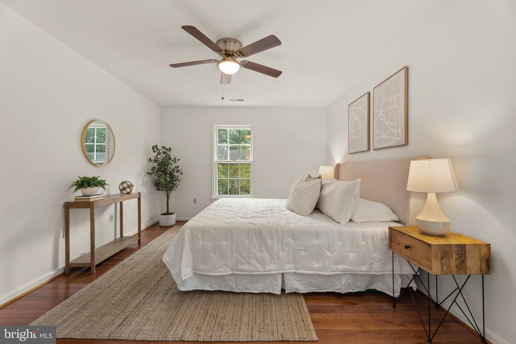 Staged bedroom with a white bed, woven rug, hardwood floors, and a window overlooking greenery.