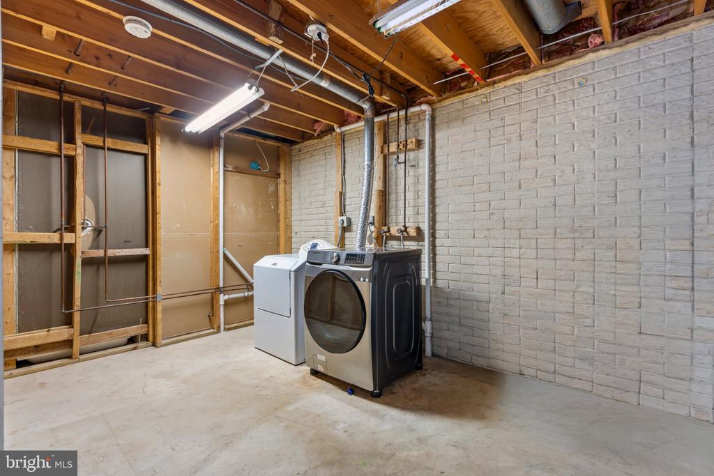 Basement laundry room with washer and dryer beside exposed framing and a white-painted brick wall.