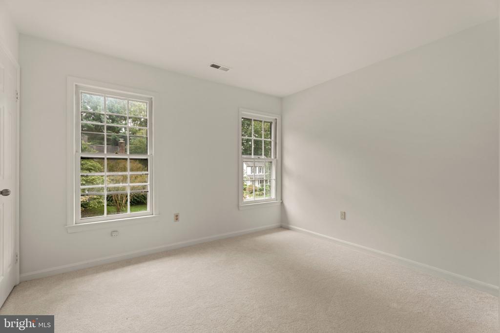 Empty bedroom with light carpet and two windows overlooking greenery.
