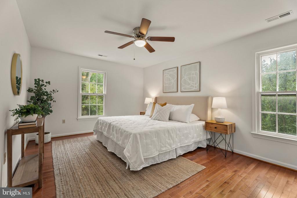 Staged bedroom with a king-size bed, white bedding, hardwood floors, and two windows overlooking trees.