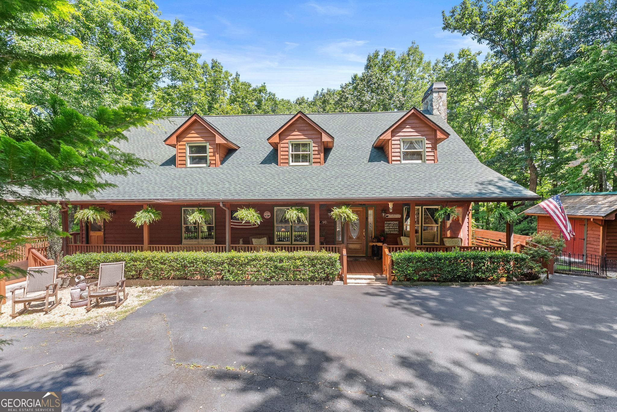 Wood-sided home with three dormers, a broad covered porch, and trees surrounding the driveway.