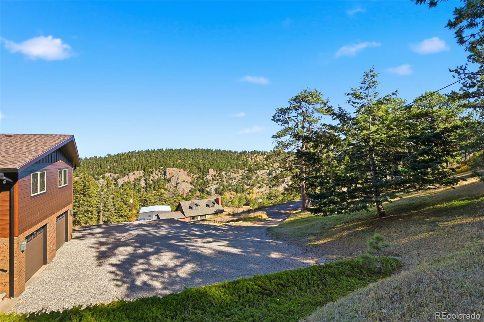 Mountain property with a gravel driveway, partial brown house, and forested rocky hills in the distance.