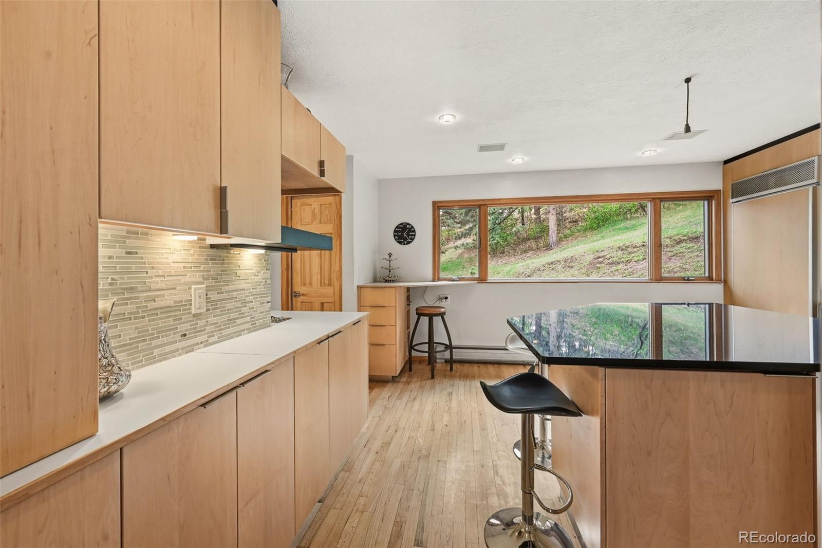 Kitchen with light wood cabinetry, a black-topped breakfast bar, and large windows overlooking a wooded hillside.