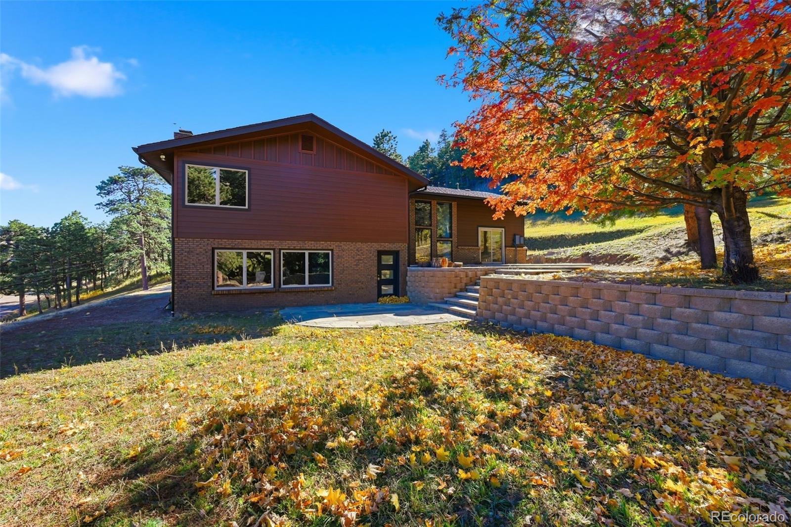 Brown-sided house with a raised patio and retaining wall beneath a large autumn tree.