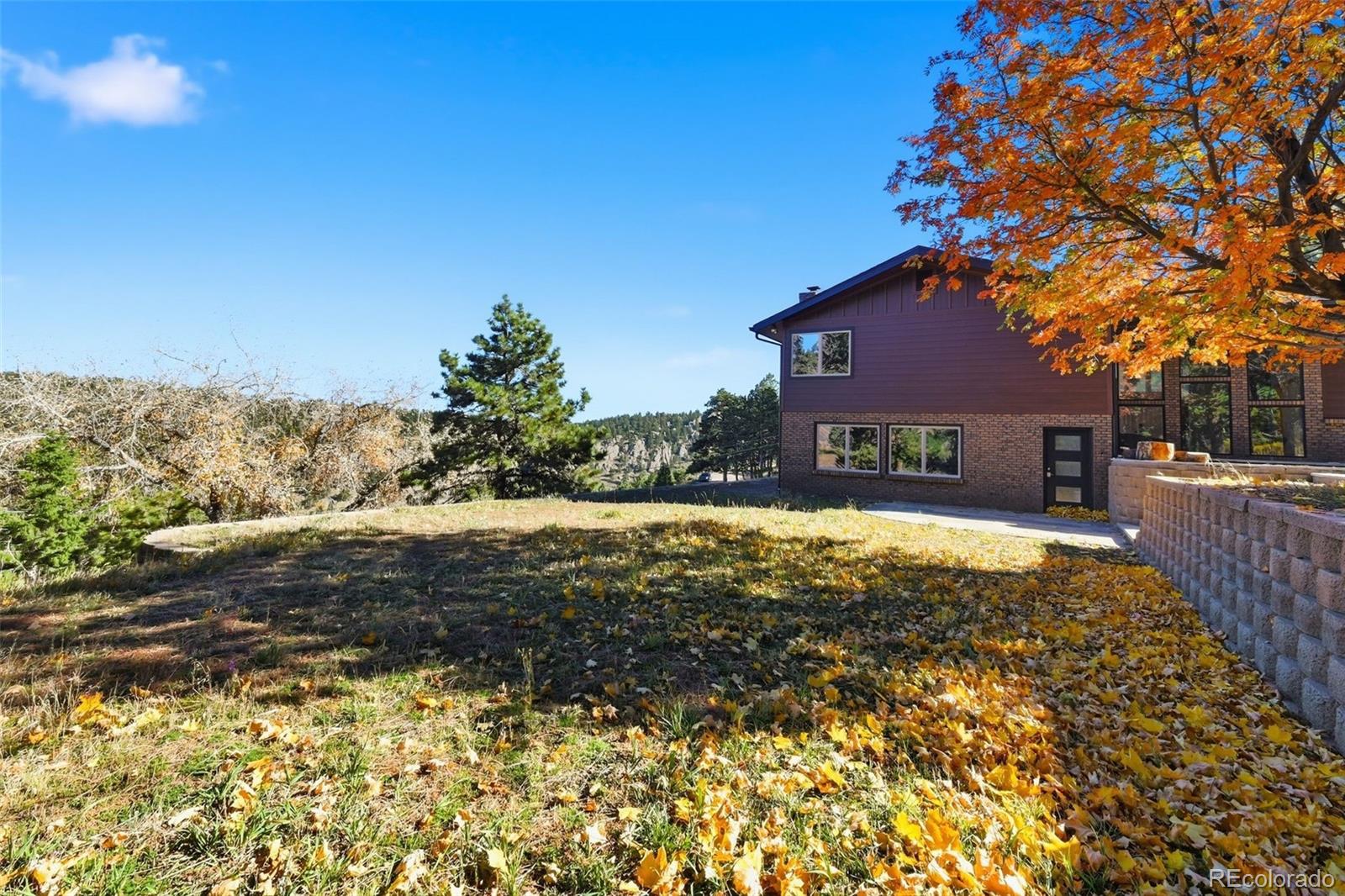 Two-story brick and dark-sided home beside an autumn yard, stone retaining wall, and wooded ridge.
