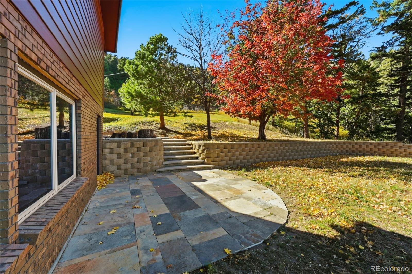 Stone patio beside a brick home, with retaining walls and autumn trees.
