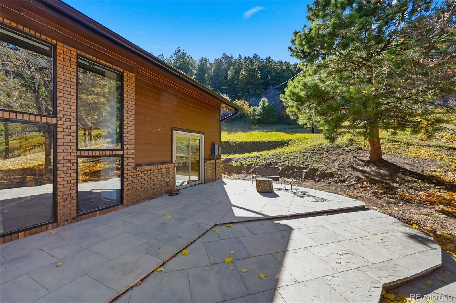 Home exterior with a broad stone patio, outdoor chairs, and a wooded hillside.