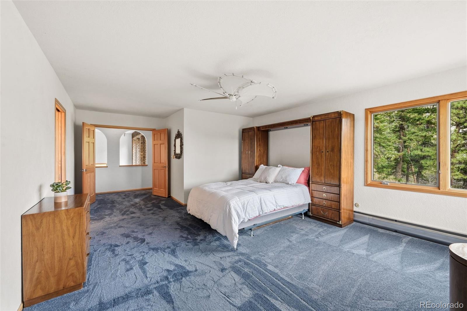 Bedroom with a white bed, blue patterned carpet, wood wardrobes, and a large window overlooking trees.