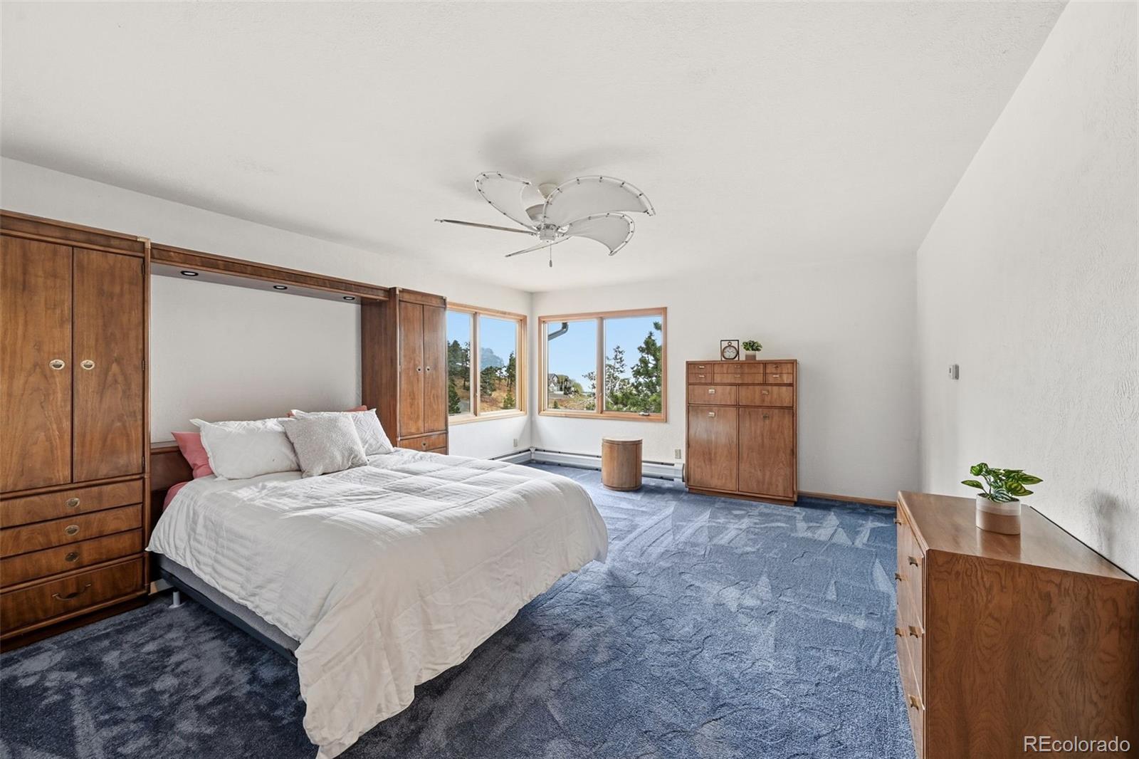 Bedroom with a white bed, blue patterned carpet, wooden wardrobes, and corner windows overlooking trees.