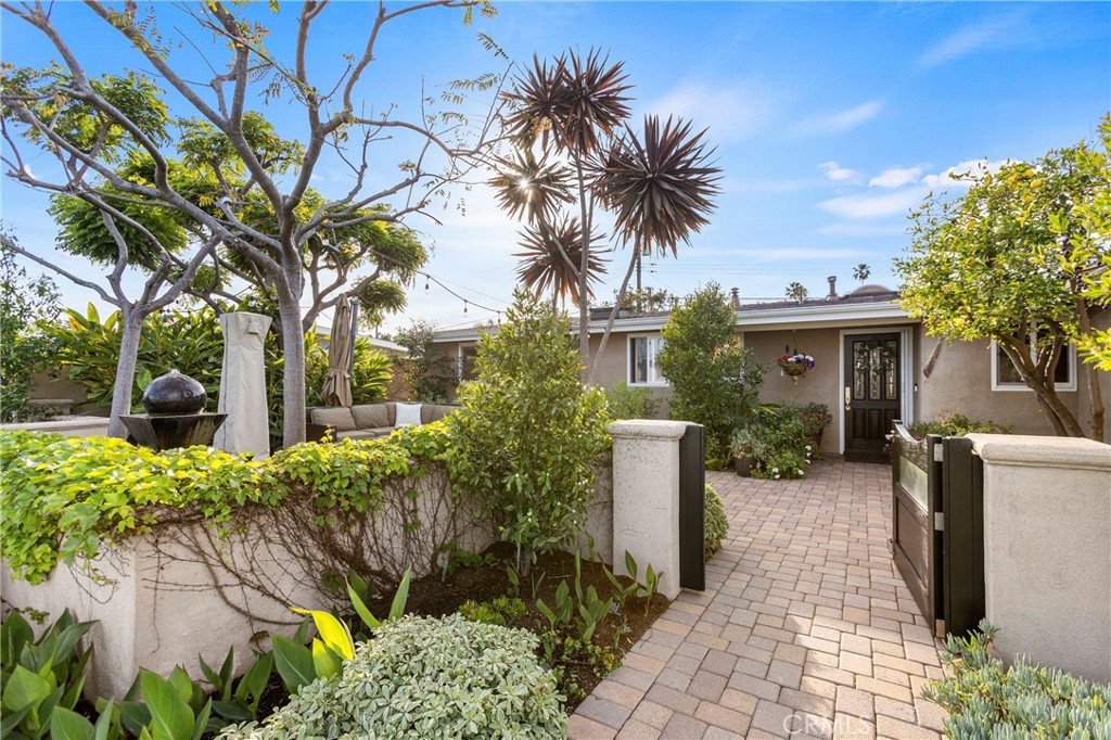 Single-story home with a paved courtyard entrance, dark front door, mature trees, and landscaped stucco walls.