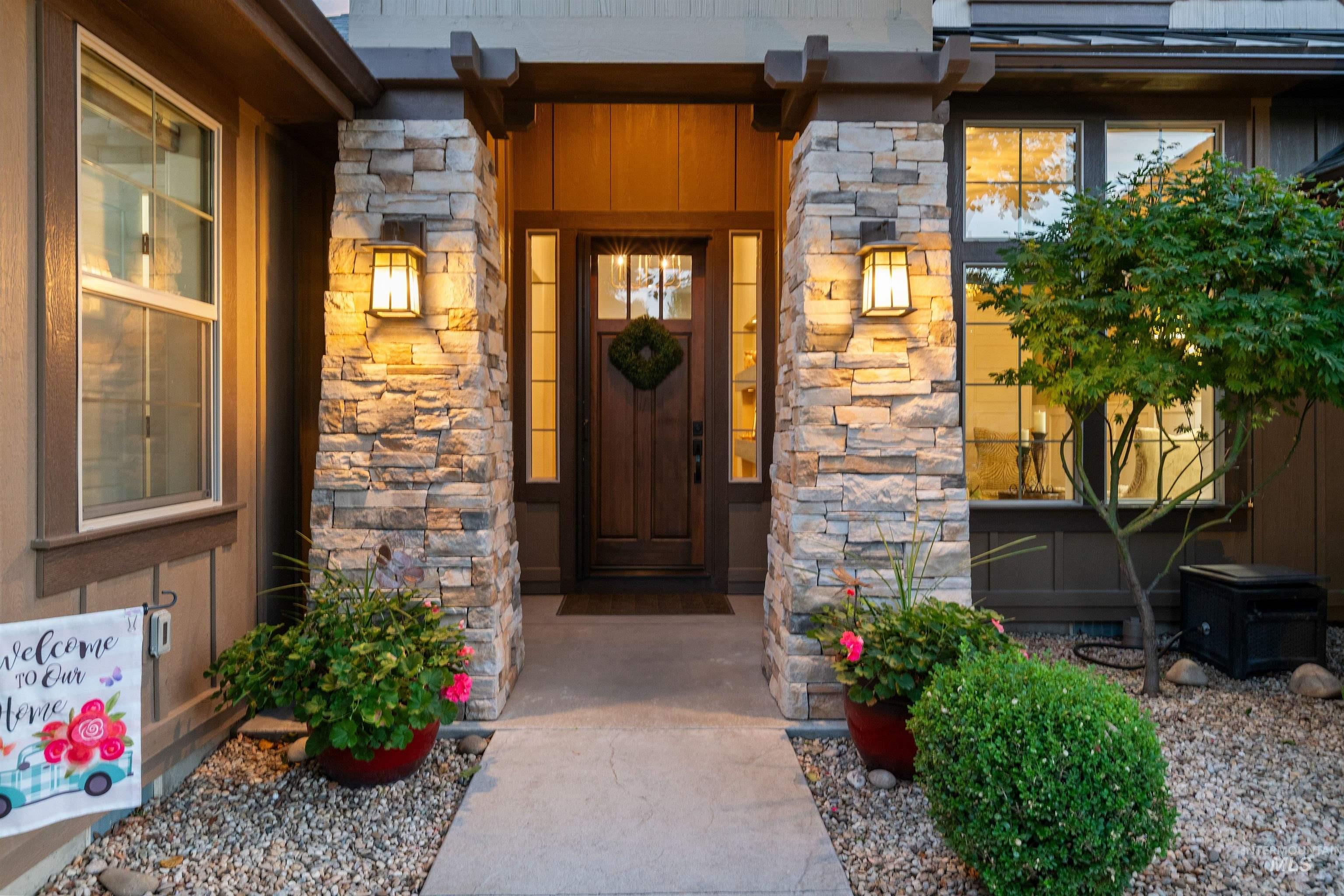 Covered front entry with stone columns, a dark wood door, glowing sconces, and flowering planters.