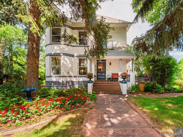 Two-story house with a covered porch, stone walkway, mature trees, and colorful flower beds.