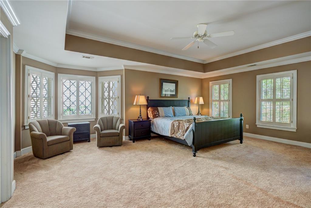 Spacious bedroom with a dark wood bed, two armchairs, beige carpet, and plantation shutters.