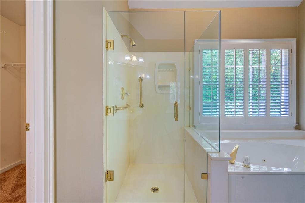 Bathroom with glass-enclosed shower, gold-tone fixtures, and a soaking tub beneath plantation shutters.