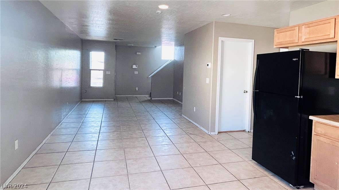 Open-plan kitchen and living area with gray tile floors, light wood cabinets, and a black refrigerator.