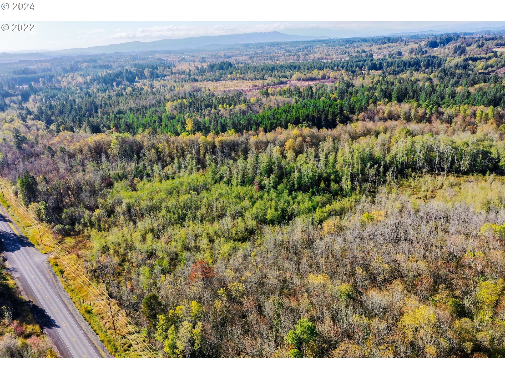 Aerial view of wooded land beside a paved road, with autumn foliage and distant hills.