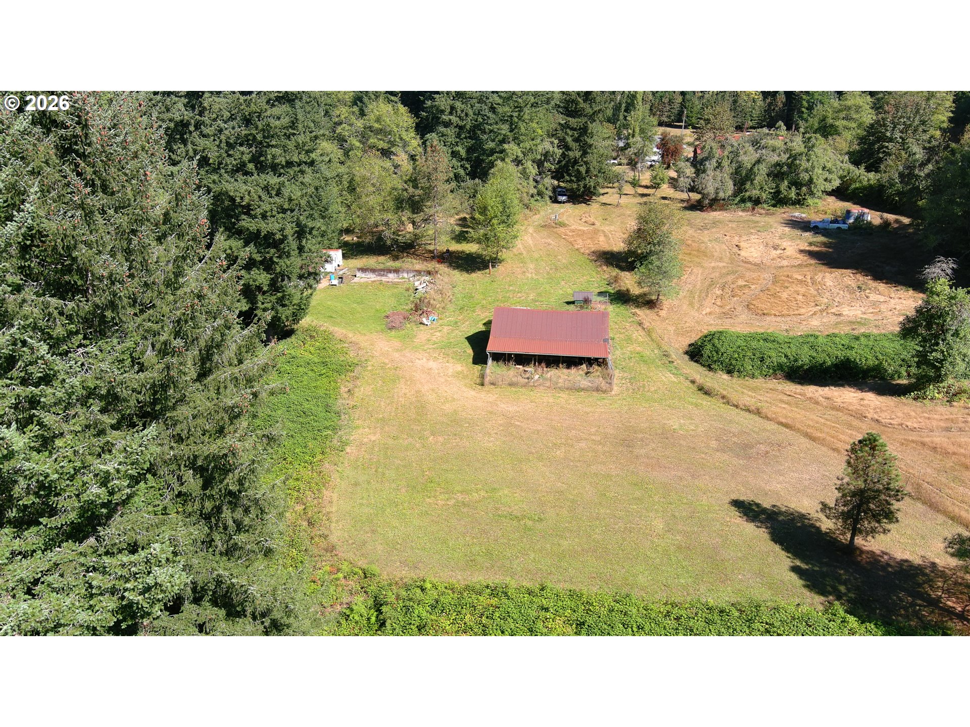 Aerial view of a grassy rural lot bordered by trees, with a small red-roofed outbuilding.