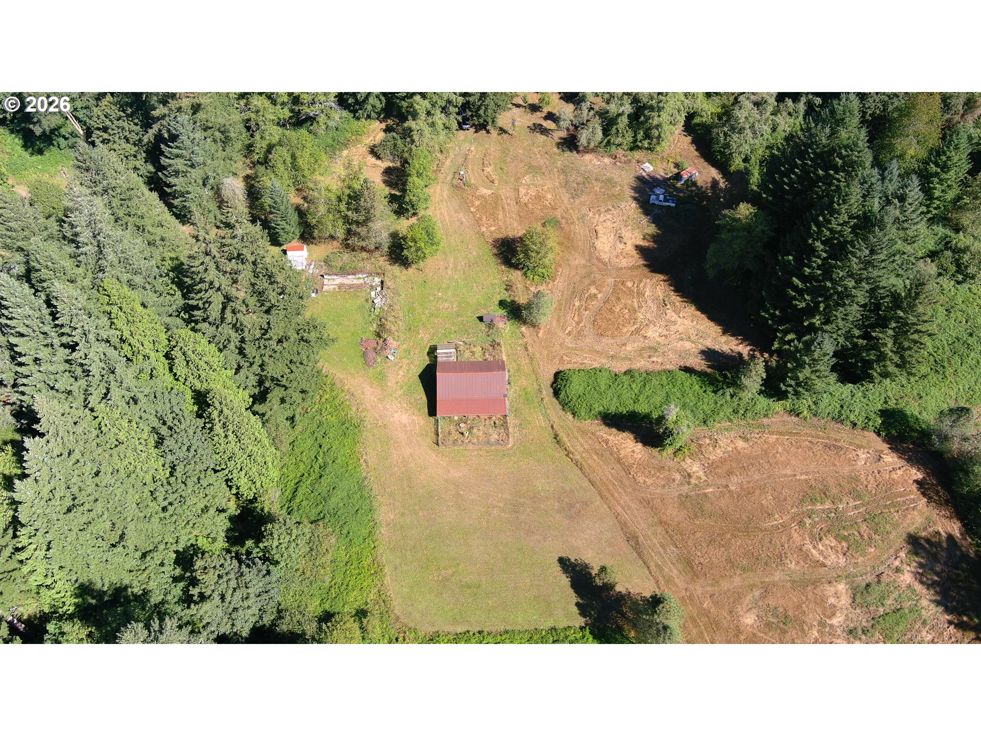 Aerial view of a wooded property with a red-roofed outbuilding and broad cleared areas.