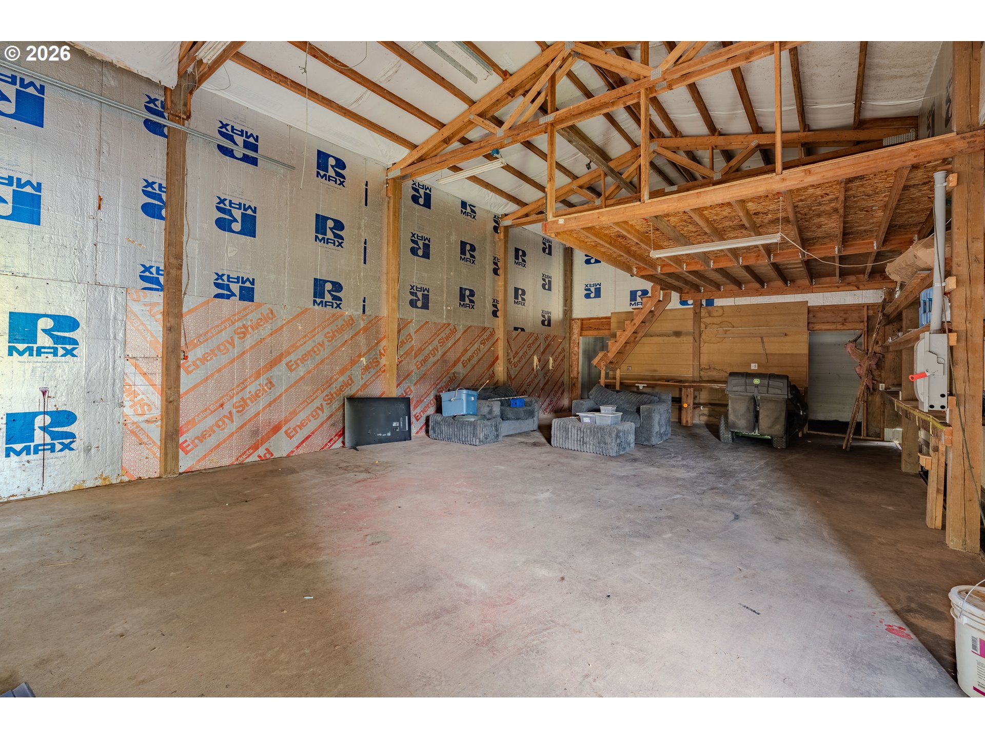 Large unfinished garage with exposed roof trusses, insulated walls, and furniture and equipment stored at the back.