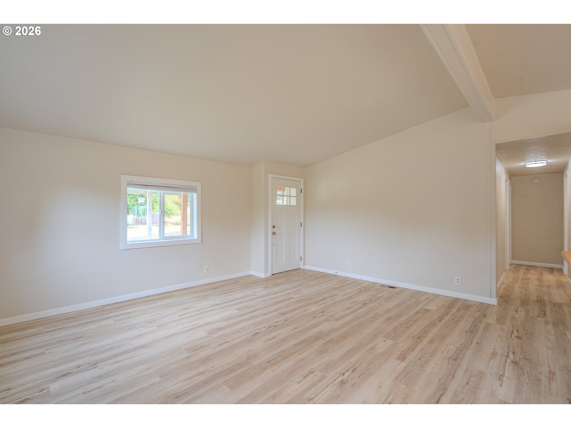 Empty living room with light wood-look flooring, a front door, and a hallway opening to the right.