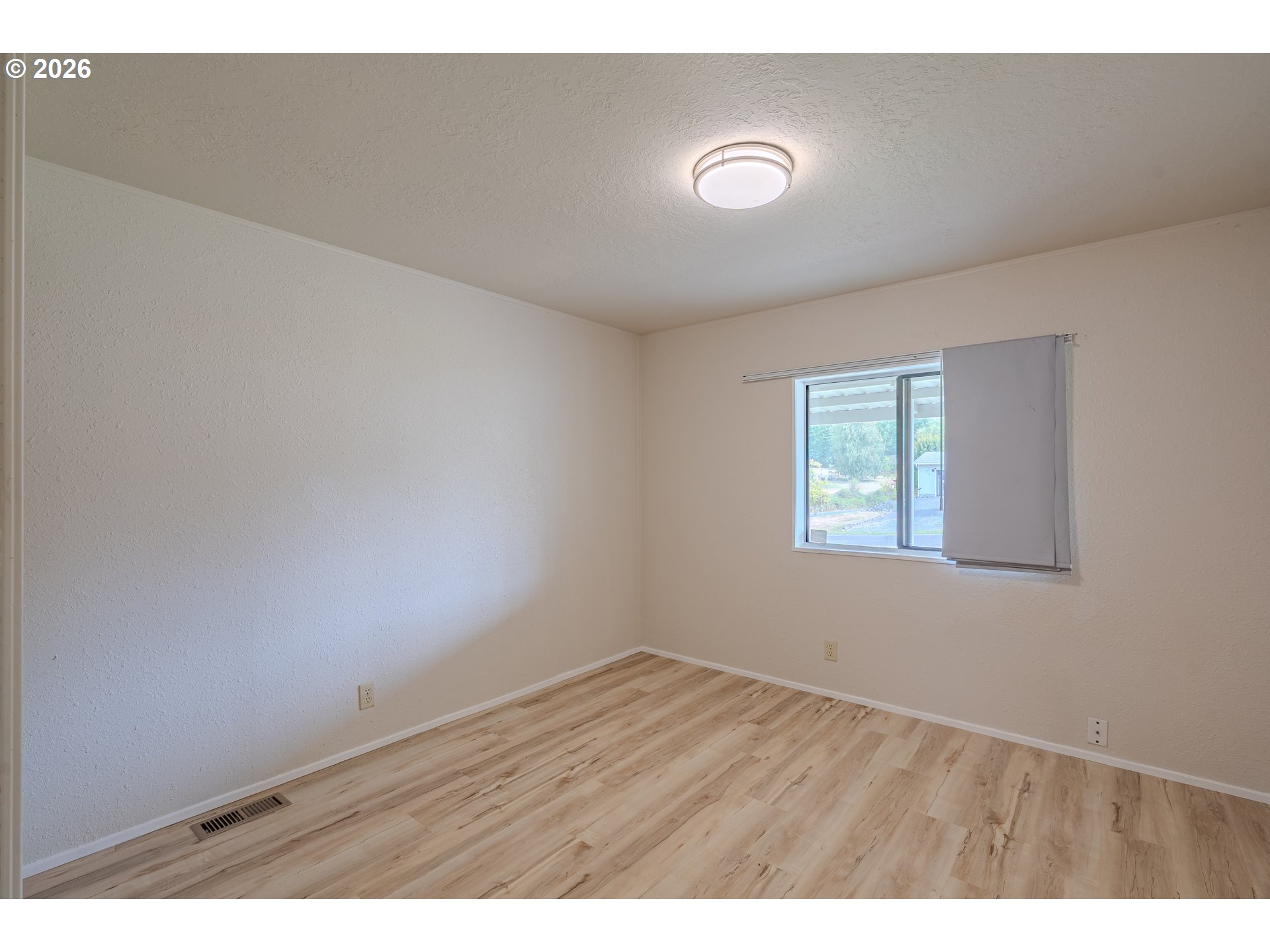Empty bedroom with light wood-look flooring and a window with a partially lowered shade.