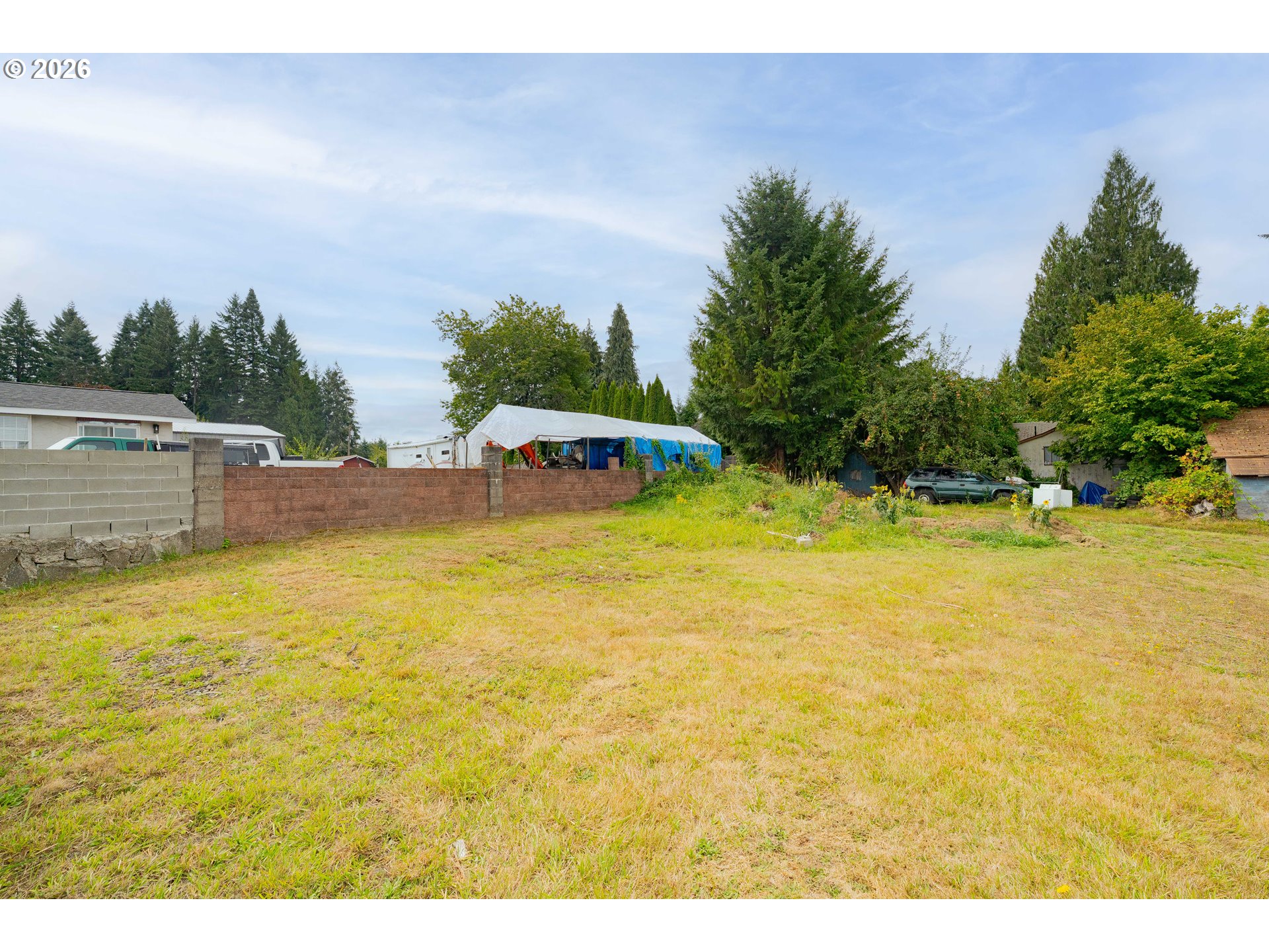 Grassy vacant lot bordered by low walls, with mature trees and neighboring homes beyond.
