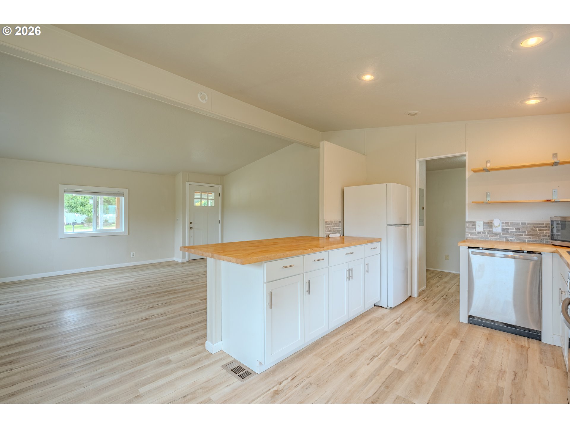 Vacant kitchen with a butcher-block island, white cabinets, and wood-look flooring opening to a living area.