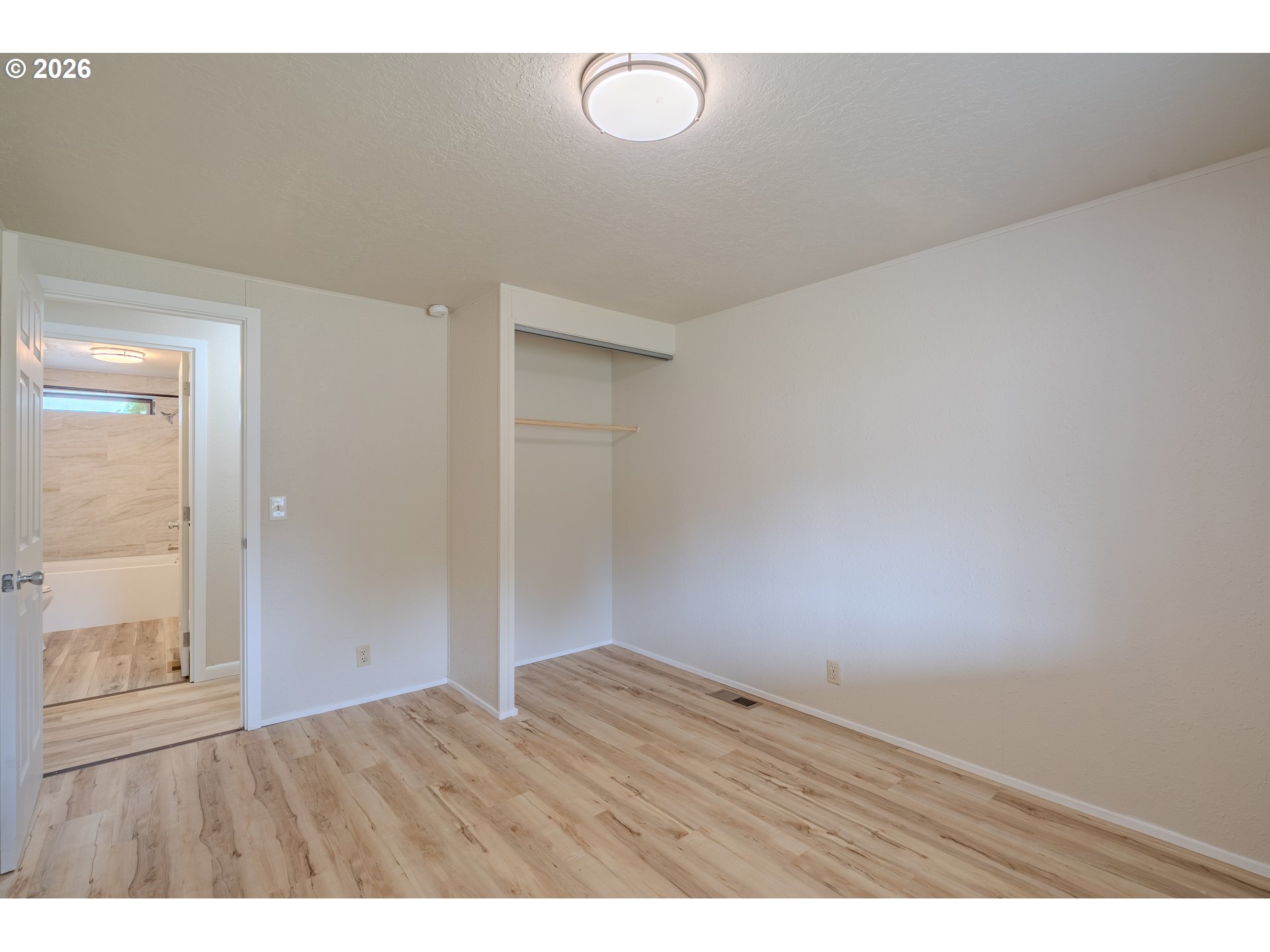 Empty bedroom with light wood-look flooring, open closet, and a bathroom visible through the doorway.