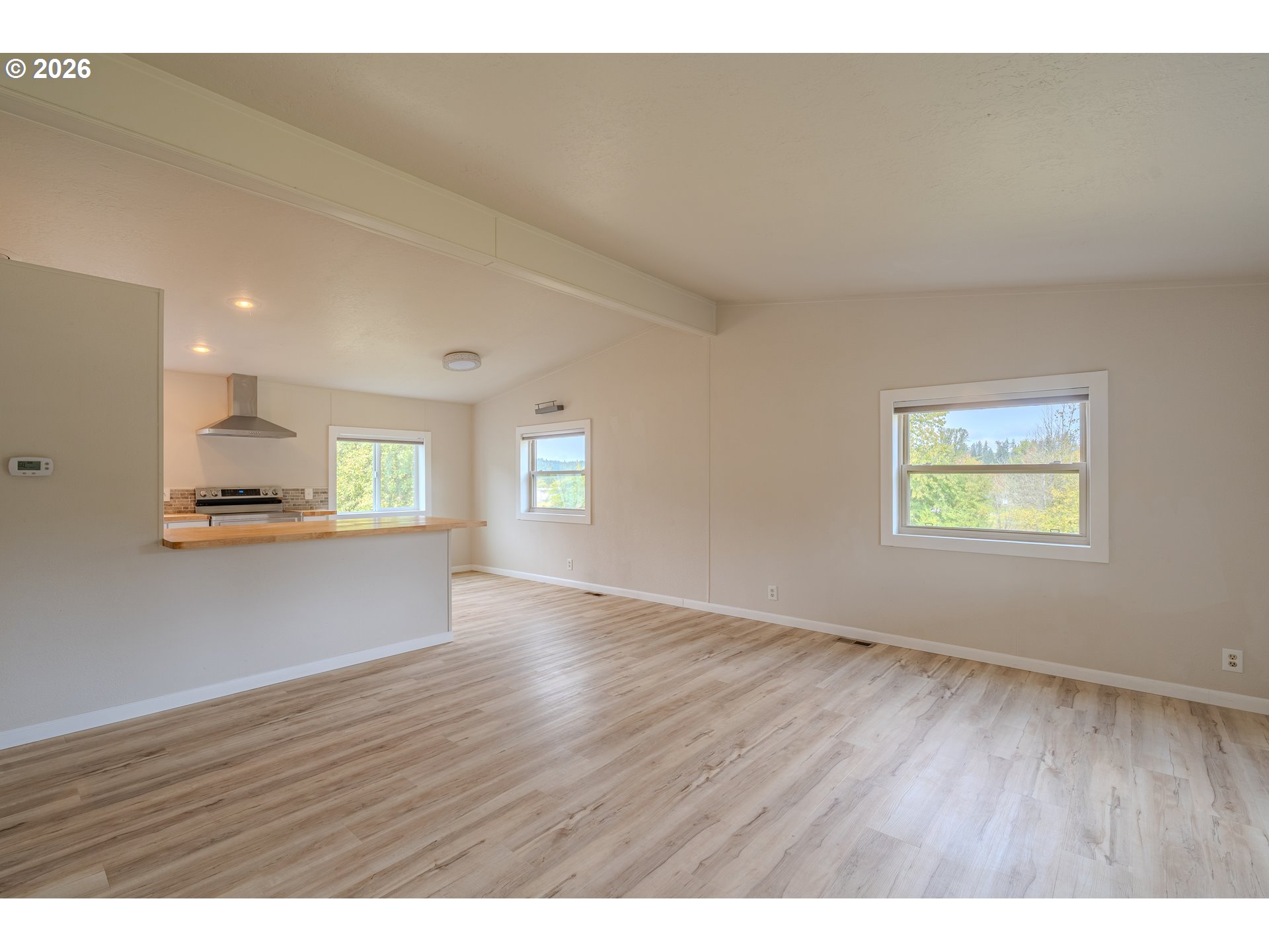 Empty living area with light wood-look floors, a breakfast bar, and windows overlooking trees.