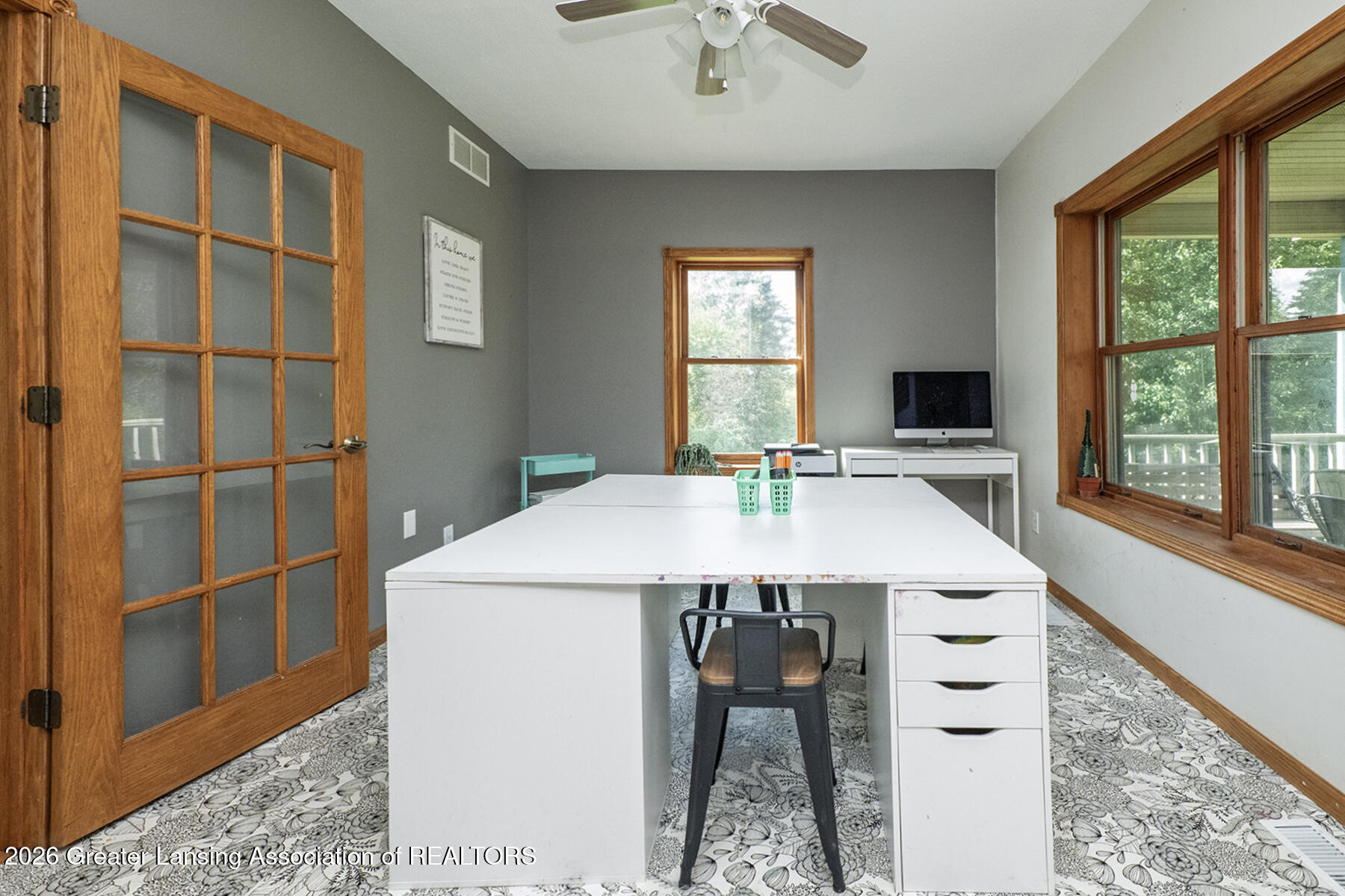 Home office with a large white desk, patterned floor covering, and wood-trimmed windows overlooking a deck.