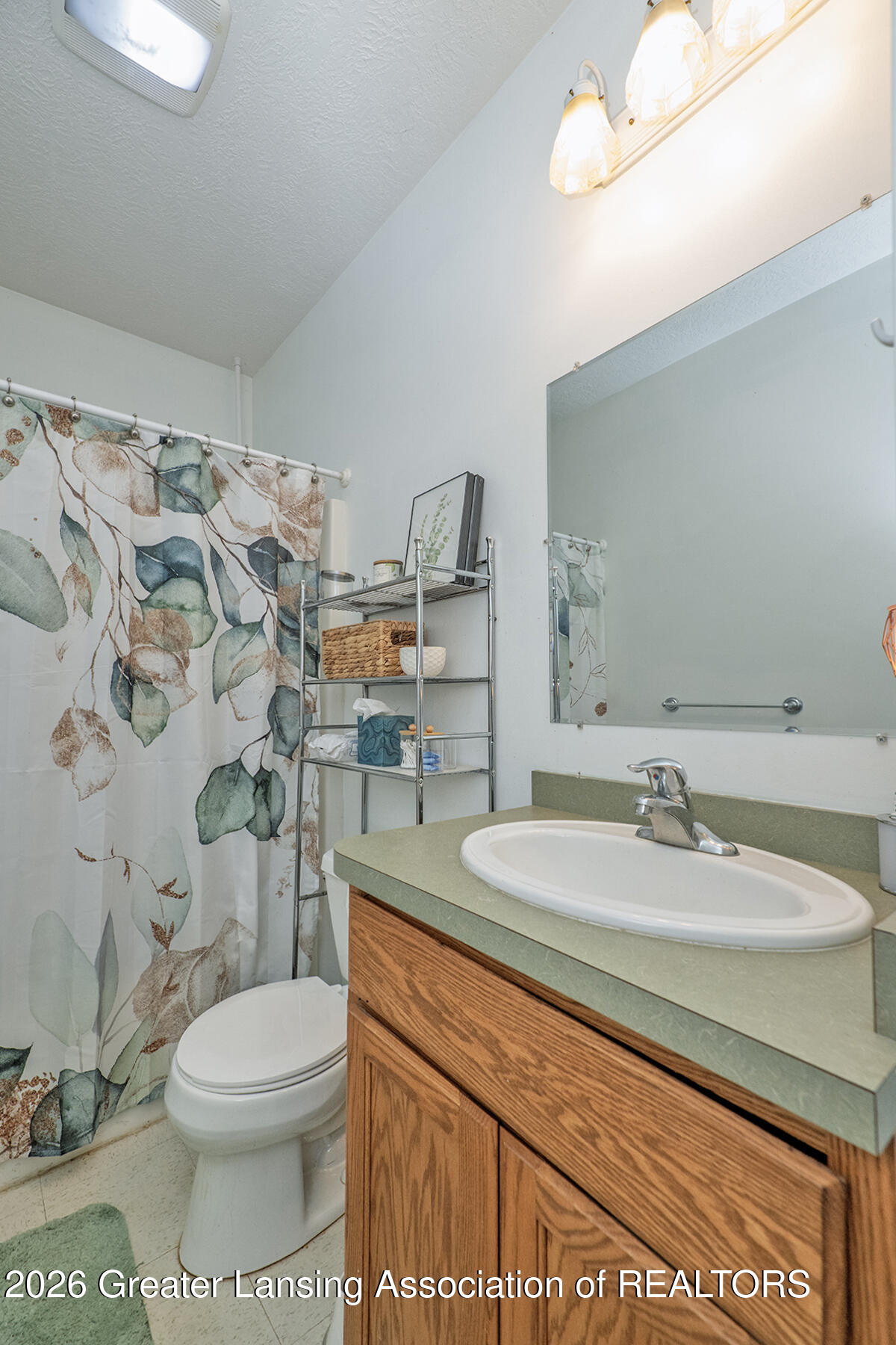 Bathroom with a wood vanity, green countertop, large mirror, toilet, and leafy-patterned shower curtain.