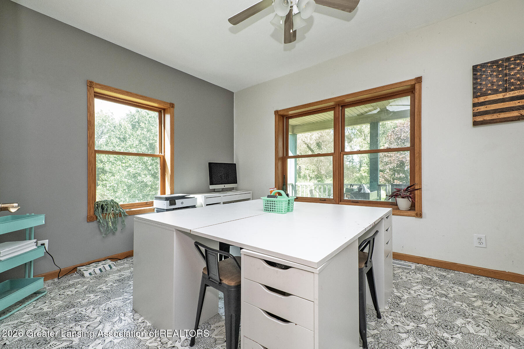 Home office with a large white desk, wood-trimmed windows, and views of trees and a covered porch.