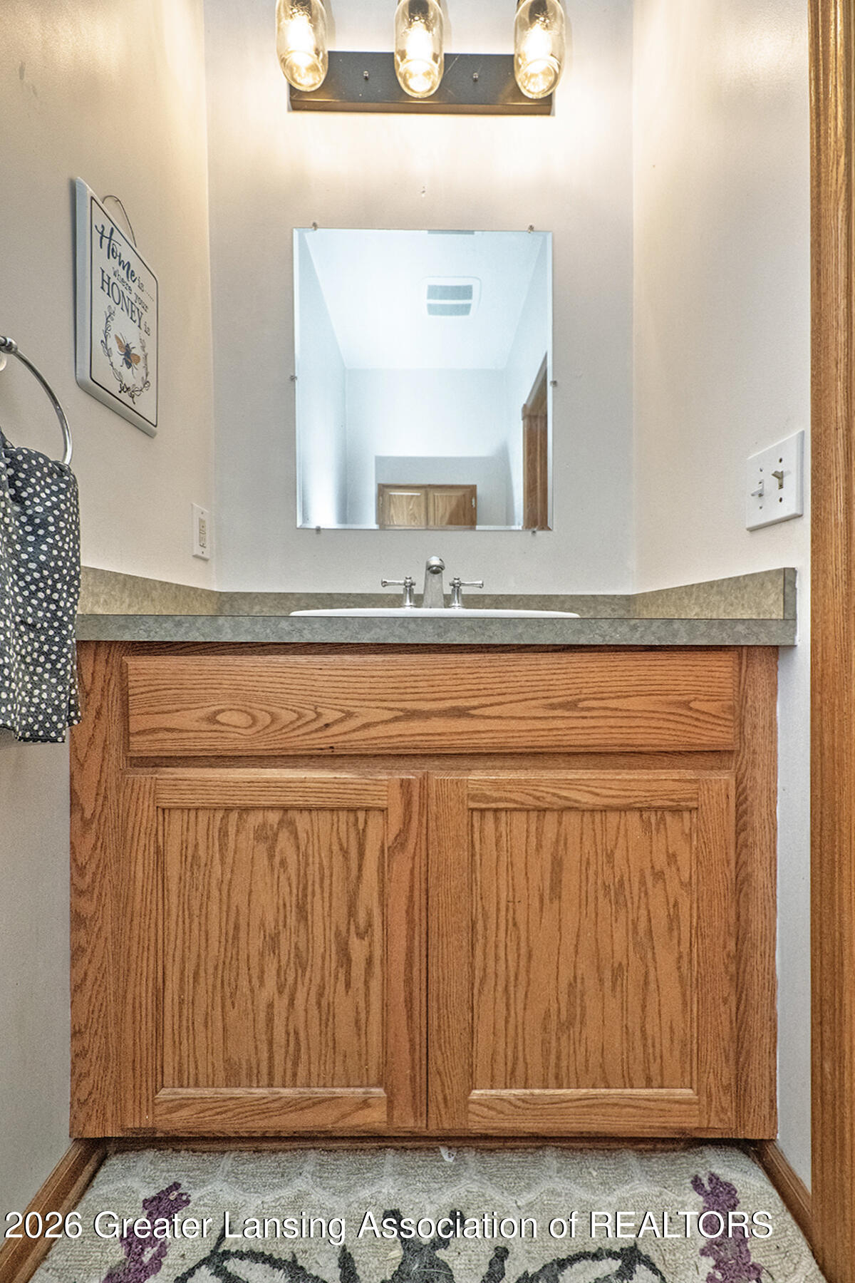 Bathroom vanity with oak cabinetry, gray countertop, rectangular mirror, and three-light fixture.