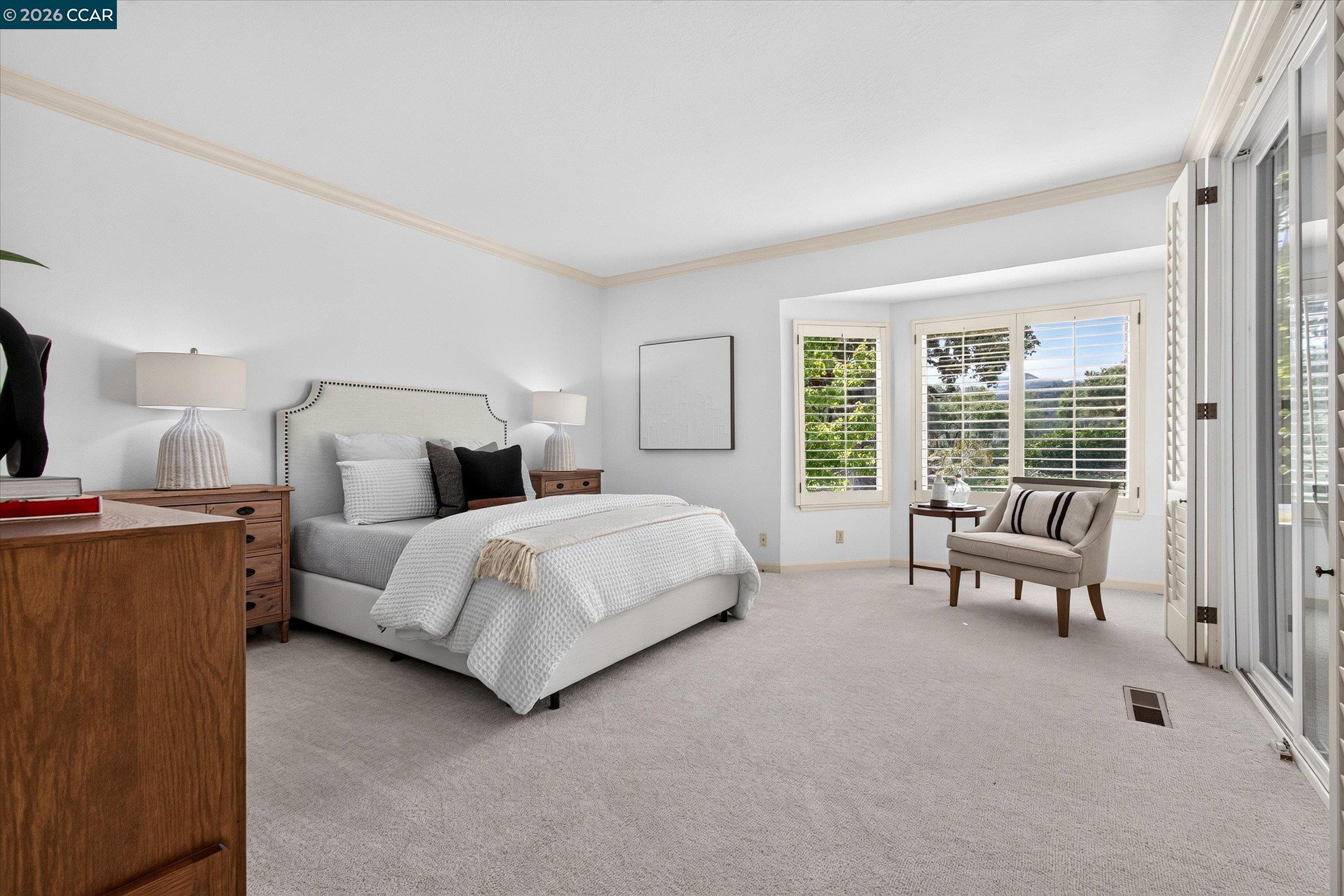Furnished bedroom with a white bed, wooden dressers, shuttered windows, and a cushioned sitting chair.