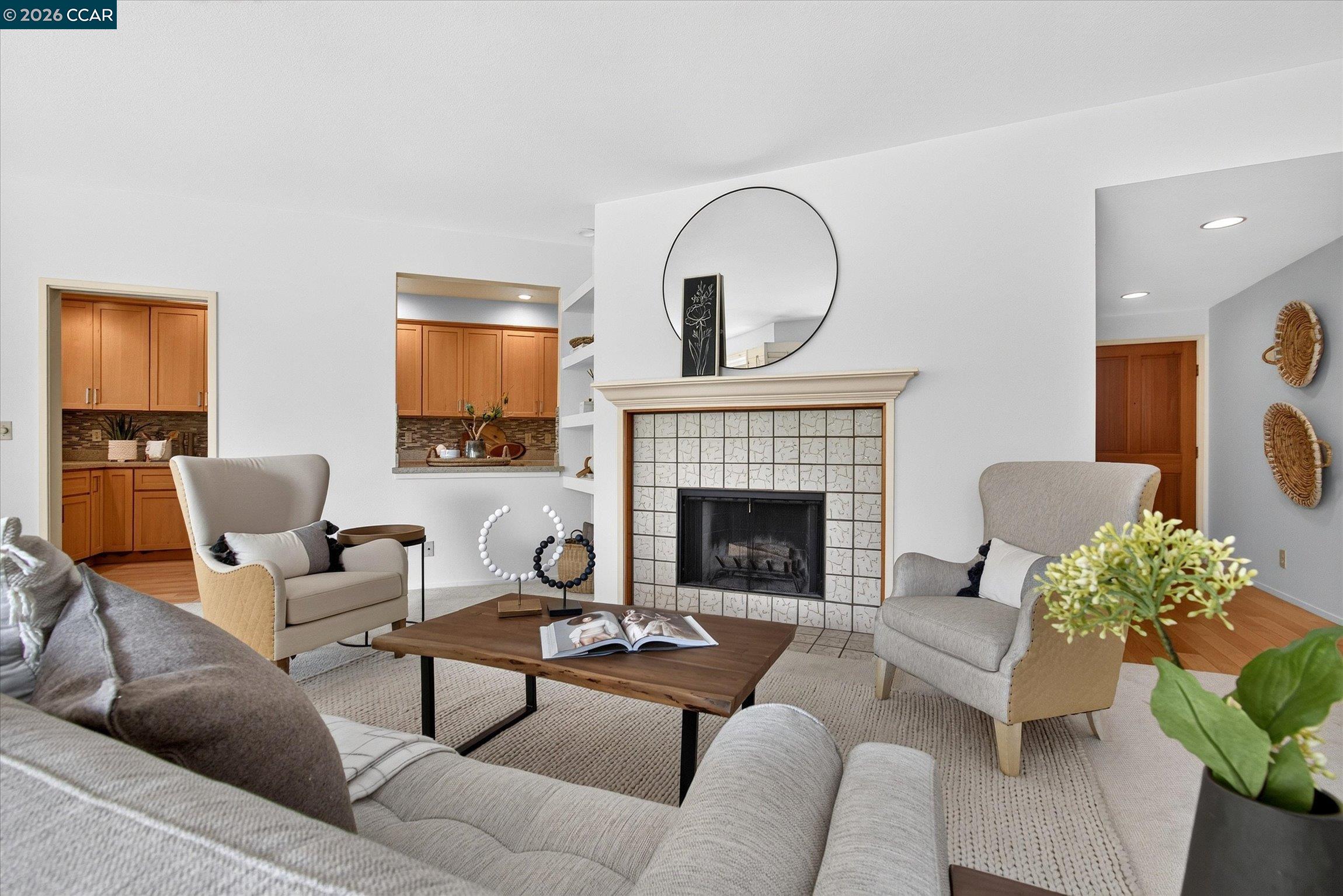 Staged living room with a tiled fireplace, round mirror, and view into the kitchen.