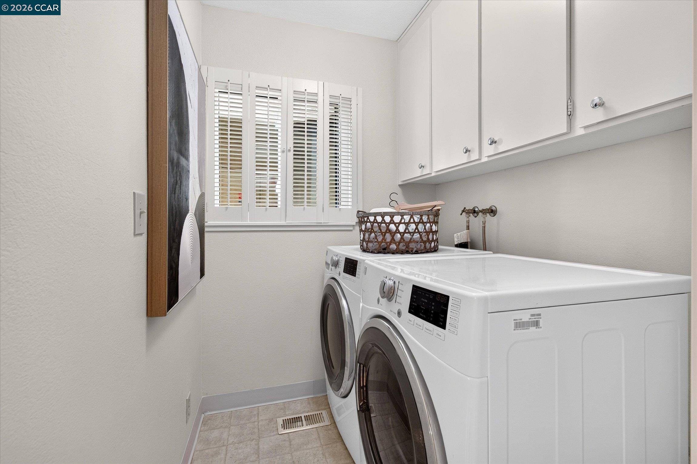 Laundry room with front-loading washer and dryer beneath cabinets, beside a shuttered window.